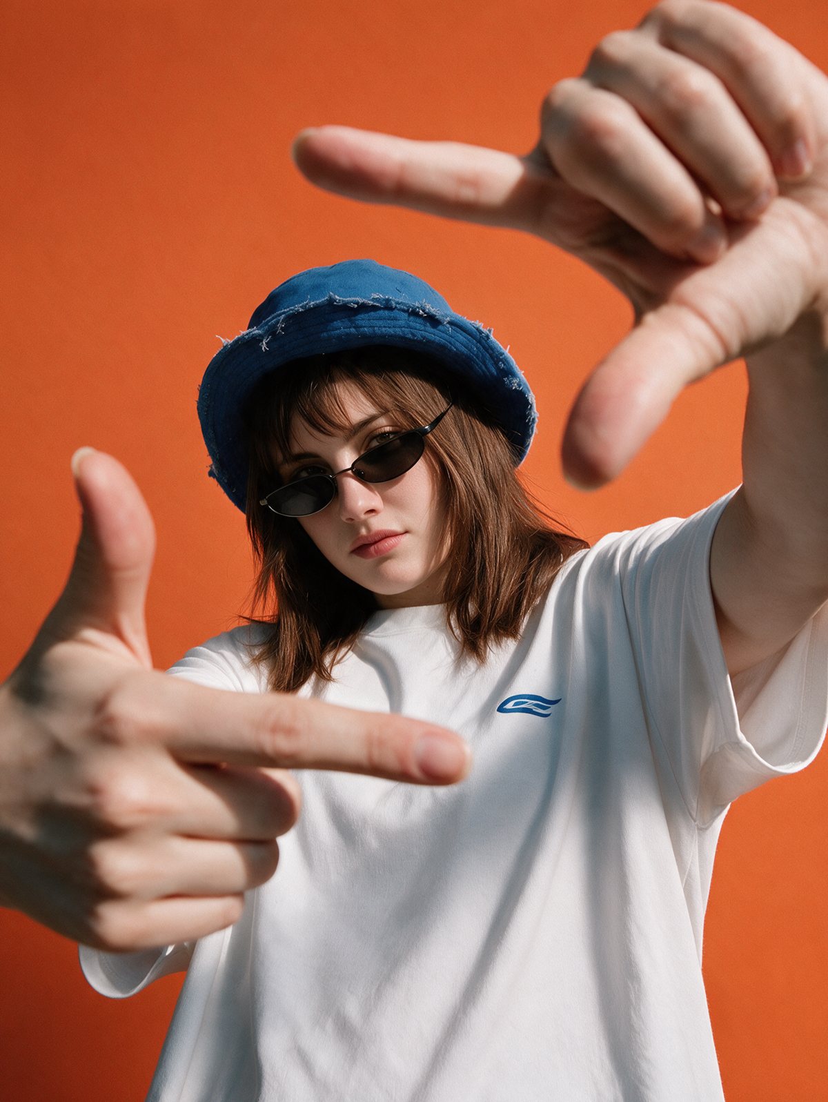 Yael con gorro azul y lentes de sol, enmarcando la cámara con las manos sobre fondo naranjo